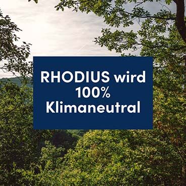 Blick in eine Waldlandschaft. In der Mitte ist eine blaue Fläche zu sehen, auf der in weißer Schrift “RHODIUS wird 100% Klimaneutral” steht.
